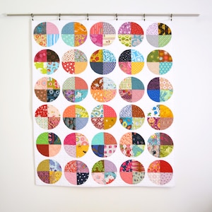May include: A colorful quilt with a white background and a pattern of large circles. Each circle is divided into four sections, each with a different patterned fabric in various colors, including pink, blue, green, and orange. The quilt is hanging from a metal rod.
