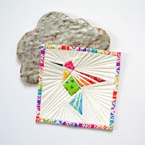 May include: A square, white quilted textile with a colorful hummingbird design. The hummingbird has a pink head, yellow and green body, and blue and red wings. The edges are trimmed with a rainbow patterned border. A textured, cloud-shaped ceramic piece sits above the textile.