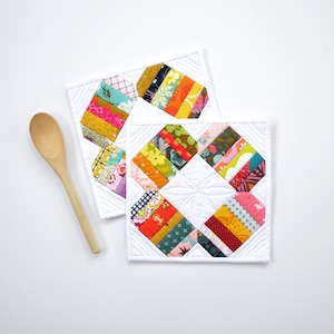 May include: Two white quilted pot holders with colorful patchwork designs. Each holder features a square shape with a central diamond pattern and vibrant strips of fabric in various colors. A wooden spoon rests nearby.