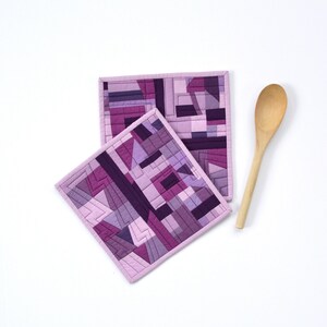 May include: Two purple geometric quilted pot holders with a light pink border. The pot holders are square and feature a patchwork design in shades of purple. A wooden spoon is next to the pot holders.