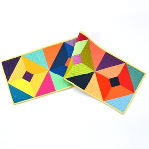 May include: Two rectangular quilted placemats featuring a vibrant geometric pattern of triangles and squares in shades of yellow, orange, red, purple, blue, teal, and pink. Each piece is finished with a bright yellow fabric binding edge.