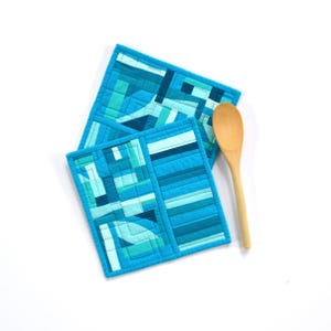 May include: Two square quilted pot holders featuring a geometric patchwork design in various shades of blue, teal, and turquoise, accompanied by a single light-colored wooden spoon placed alongside the fabric items on a plain white surface.