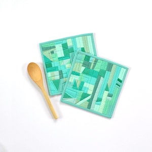 May include: Two square quilted potholders featuring a geometric patchwork design in various shades of green, including mint, emerald, and lime, paired with a single light-colored wooden spoon with a smooth, rounded handle and a deep oval bowl.
