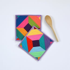 May include: Two square, quilted pot holders with a geometric design in vibrant colors including pink, orange, green, and purple. A wooden spoon rests nearby. The pot holders have a purple border.