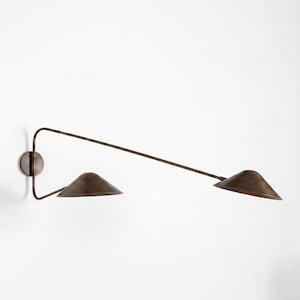 Könnte beinhalten: Bronze Wandleuchte mit zwei konischen Schirmen. Die Leuchte hat einen kreisförmigen Sockel und einen langen, abgewinkelten Arm, der sich zu den Schirmen erstreckt. Die Schirme sind in verschiedenen Winkeln positioniert, was ein modernes Design schafft. Die Leuchte ist an einer weißen Wand montiert.