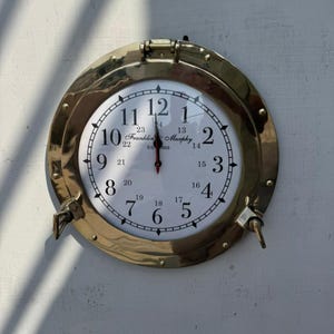 Könnte beinhalten: Eine runde Messing-Uhr mit weißem Zifferblatt und schwarzen Ziffern. Die Uhr hat ein nautisches Bullauge-Design mit einer Scharnierblende. Das Zifferblatt zeigt den Text "Franklin & Murphy, Est 1868". Die Zeiger zeigen auf 12:00 Uhr.