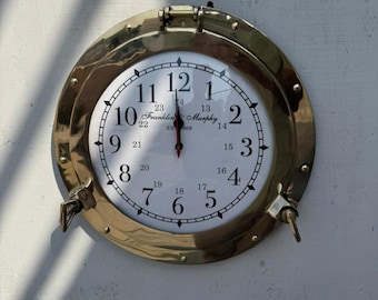 May include: A round brass clock with a white face and black numerals. The clock has a nautical porthole design with a hinged bezel. The clock face displays the text "Franklin & Murphy, Est 1868". The hands point to 12:00.