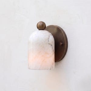 Può includere: Applique da parete con paralume effetto marmo che emette una luce calda e diffusa. Il paralume ha una forma a campana arrotondata, sormontata da una piccola sfera color bronzo. Montato su una piastra posteriore circolare color bronzo.