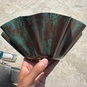 Op de afbeelding: Een gegroefde, waaiervormige kom met een donkerbruine basiskleur en een verweerde, groenblauwe patina-afwerking. De kom heeft een brede, open bovenkant met diepe, verticale ribbels die vanuit het midden uitstralen en een rand vormen.
