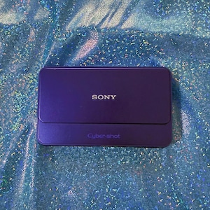 May include: A rectangular purple Sony Cyber-shot digital camera with a smooth, matte finish. The word SONY is printed in white on the upper section, and the text Cyber-shot is printed in a darker purple on the lower section of the device.
