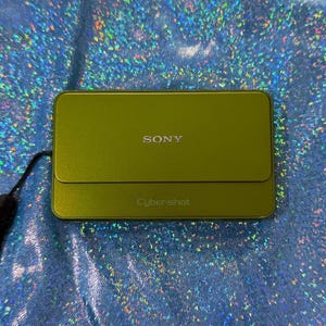 May include: A rectangular olive green Sony Cyber-shot digital camera with a sliding lens cover. The word SONY is printed in silver on the upper portion, and the text Cyber-shot is embossed on the lower portion. A black wrist strap is attached.