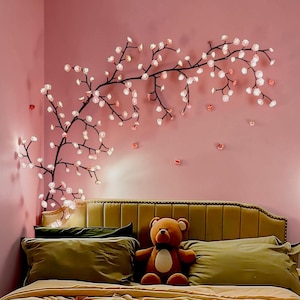 May include: A wall decoration featuring a black branch design with illuminated white and pink blossoms. The art is mounted on a pink wall above a bed with olive green pillows and a brown teddy bear.