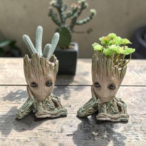 Puede incluir: Dos macetas con forma de Groot, una con un cactus y otra con suculentas verdes. Las macetas son marrones con una textura de madera y tienen una cara sonriente. Las plantas están frente a un fondo borroso.