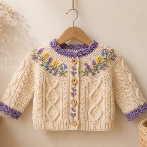 May include: A cream-colored crocheted cardigan featuring a cable knit pattern, long sleeves, and a round neckline. The garment is accented with purple scalloped trim and embroidered floral details in yellow, blue, and purple, fastened with five wooden buttons.