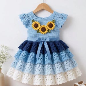 May include: A light blue crochet dress featuring three yellow sunflower appliqués with brown centers on the bodice. The skirt consists of four tiered layers in shades of dark blue, medium blue, light blue, and white, accented by a blue satin ribbon bow.