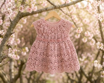 May include: A dusty pink crochet dress featuring an intricate openwork pattern, a round neckline, and short sleeves. The bodice is adorned with three crocheted floral embellishments on the upper right side and a matching satin ribbon at the waist.