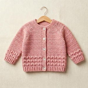 Può includere: Un cardigan da bambino all'uncinetto rosa chiaro con maniche lunghe e scollo tondo. Il maglione con bottoni presenta cuciture decorative sul fondo e sui polsini delle maniche ed è esposto su una gruccia di legno.