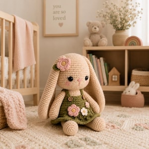 May include: A beige crochet bunny doll with long ears, wearing an olive green dress adorned with small pink and white crocheted flowers and two brown buttons. The bunny has black eyes, pink cheeks, and a small pink stitched nose, sitting upright.