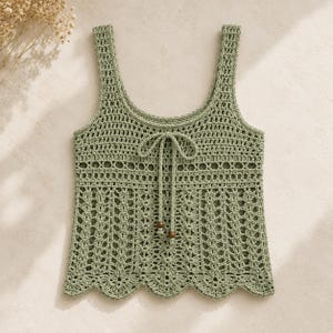 May include: A sage green sleeveless crochet tank top featuring a scoop neckline, a drawstring tie with two small wooden beads at the ends, and a scalloped hem. The garment displays a combination of openwork patterns and vertical stitch textures.