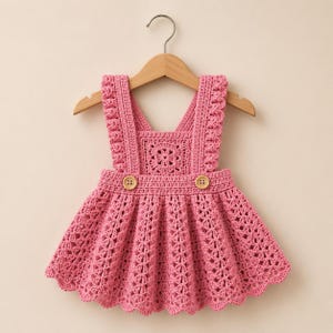 Könnte beinhalten: Ein rosa Häkel-Trägerkleid mit quadratischem Muster am Oberteil, breiten Schulterträgern mit strukturierten Kanten und einem ausgestellten Rock mit gewelltem Saum. Zwei hellbraune Holzknöpfe sind an der vorderen Taille angebracht.