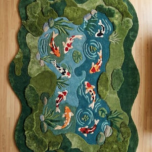 May include: A unique, handcrafted rug depicting a koi pond scene. The rug features a blue pond with orange, white, and black koi fish, surrounded by green and darker green textured areas representing foliage. The rug's shape is irregular, mirroring the pond's outline.