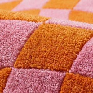 May include: A tufted textile featuring a repeating checkered pattern in vibrant orange and soft pink. The surface has a thick, plush, and textured pile construction, creating a tactile grid of alternating coloured squares across the entire fabric.