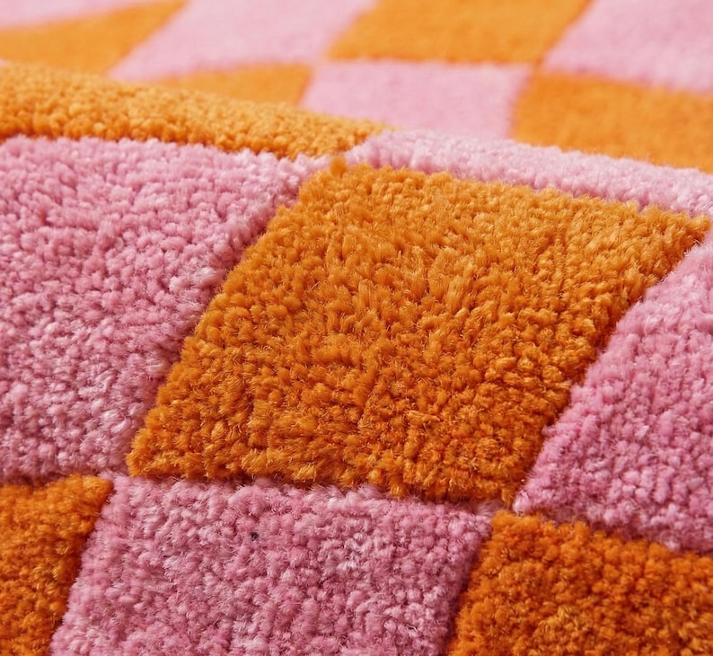 May include: A tufted textile featuring a repeating checkered pattern in vibrant orange and soft pink. The surface has a thick, plush, and textured pile construction, creating a tactile grid of alternating coloured squares across the entire fabric.