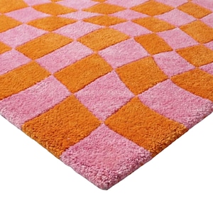 May include: A tufted rug featuring a repeating checkerboard pattern in alternating bright orange and soft pink squares. The textile surface has a dense, plush pile texture that creates a tactile appearance across the entire square-shaped mat.