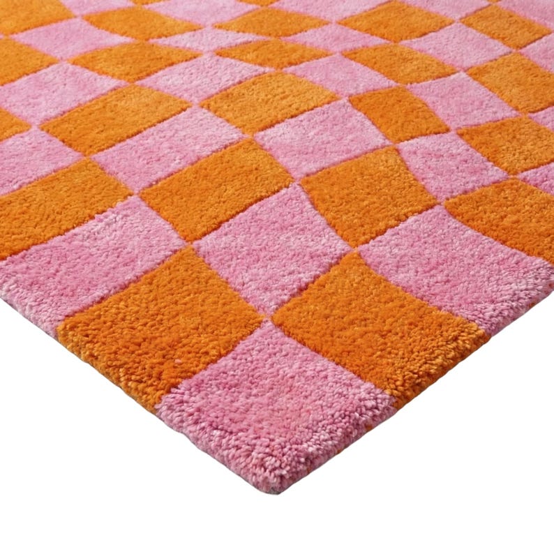 May include: A tufted rug featuring a repeating checkerboard pattern in alternating bright orange and soft pink squares. The textile surface has a dense, plush pile texture that creates a tactile appearance across the entire square-shaped mat.