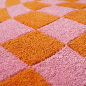 May include: A textile surface featuring a repeating checkerboard pattern of alternating bright orange and soft pink squares. The material has a dense, looped pile texture that creates a raised, tactile appearance across the entire surface area.