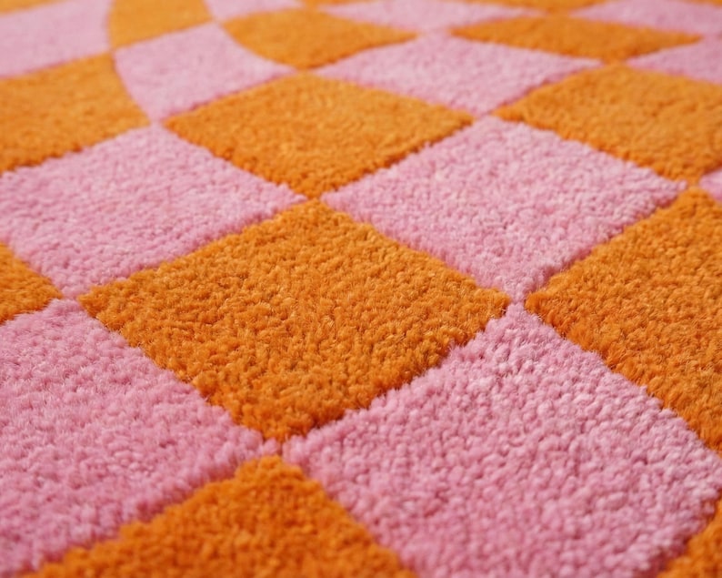 May include: A textile surface featuring a repeating checkerboard pattern of alternating bright orange and soft pink squares. The material has a dense, looped pile texture that creates a raised, tactile appearance across the entire surface area.
