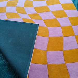 May include: A tufted rug featuring a distorted checkerboard pattern in alternating shades of mustard yellow and light lavender. The rug is partially folded over to reveal a solid dark teal fabric backing on the underside of the textile piece.