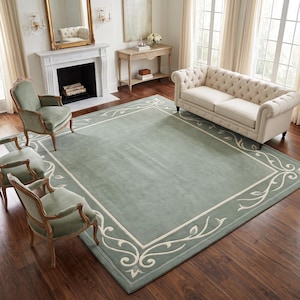 May include: A large rectangular area rug featuring a solid sage green centre field surrounded by a wide border with a cream-coloured scrolling vine and leaf pattern. The rug is positioned on a dark wood floor in a room with furniture.