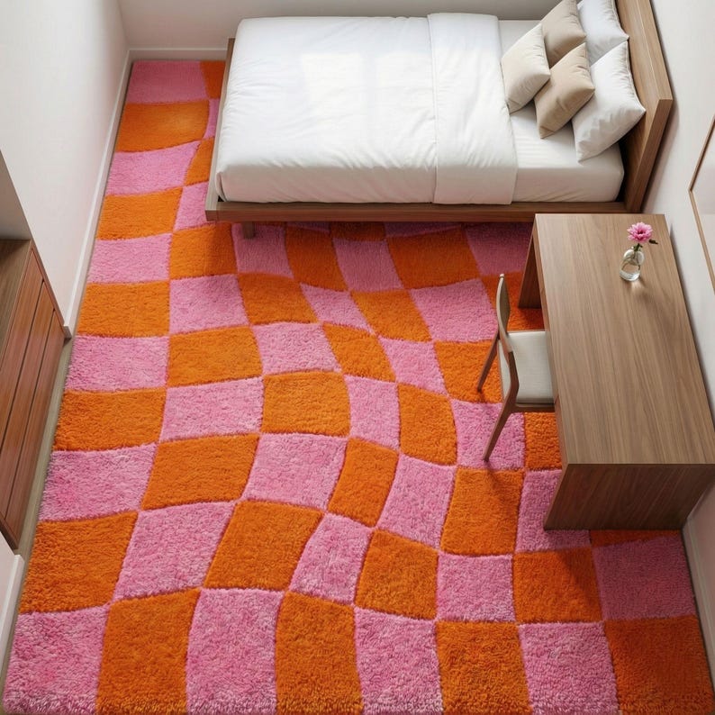 May include: A rectangular area rug featuring a vibrant, wavy checkerboard pattern in alternating shades of bright pink and orange. The rug has a thick, textured pile and covers the floor space beneath a wooden bed frame and a wooden desk.