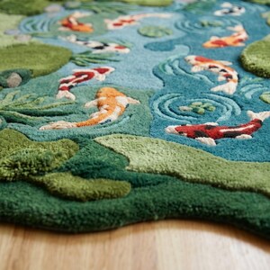 May include: A hand-tufted rug depicting a koi pond scene. The rug features a blue pond with orange, red, and black koi fish swimming among green lily pads and surrounding foliage. The rug is made of soft, textured material.