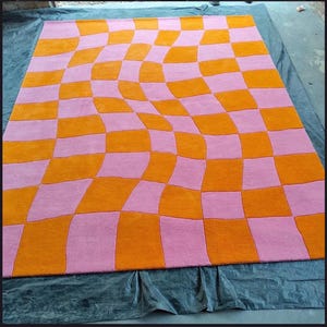 May include: A rectangular area rug featuring a distorted checkerboard pattern. The design consists of alternating squares in vibrant orange and soft pink, arranged in a wavy, warped grid that creates a sense of movement across the surface.