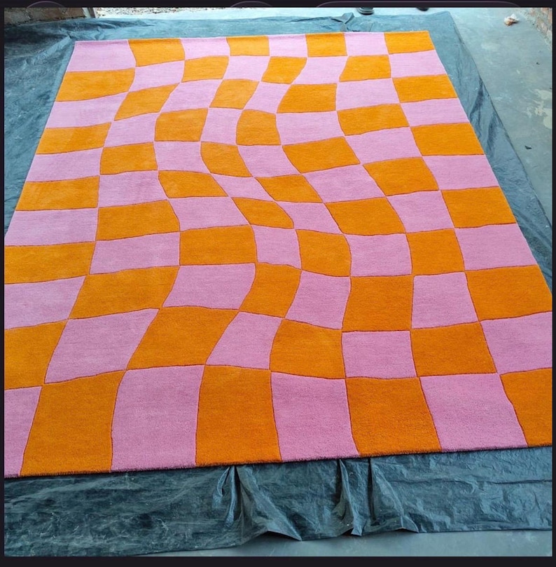 May include: A rectangular area rug featuring a distorted checkerboard pattern. The design consists of alternating squares in vibrant orange and soft pink, arranged in a wavy, warped grid that creates a sense of movement across the surface.