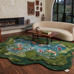May include: A unique, handcrafted rug shaped like a pond, featuring koi fish in shades of orange, white, and black. The rug has a green border with textured details, creating a 3D effect. The rug is placed in a living room setting.