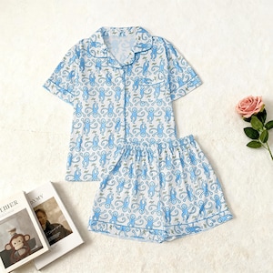 May include: A light blue and white pajama set featuring a playful monkey pattern. The set includes a short-sleeved button-down shirt and matching shorts. The edges of the shirt and shorts are trimmed with a light blue border.