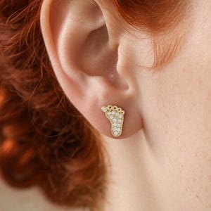 May include: A gold-colored stud earring in the shape of a baby footprint, encrusted with small, clear gemstones. The earring is worn on the ear of a person with red hair.