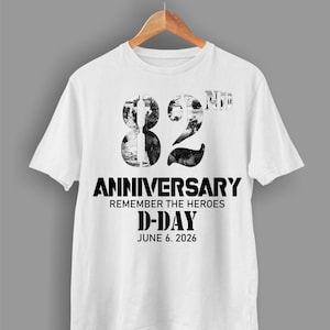 Puede incluir: Camiseta blanca con un gráfico en blanco y negro que conmemora el 82º aniversario del Día D. El diseño incluye el número "82" relleno con una imagen en escala de grises, la palabra "ANIVERSARIO" y el texto "REMEMBER THE HEROES D-DAY JUNE 6. 2026".