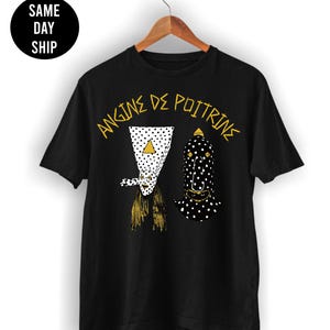 May include: A black short-sleeved t-shirt featuring a graphic print of two figures with polka-dot patterns and the yellow text "ANGINE DE POITRINE" arched above them. The shirt is displayed on a wooden hanger against a plain white background.