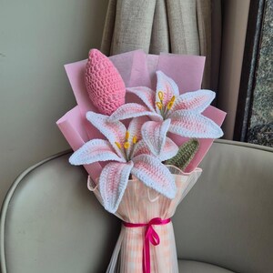 Könnte beinhalten: Ein handgefertigter Häkelblumenstrauß mit zwei großen weißen und rosafarbenen Lilien mit gelben Staubgefäßen, einer einzelnen rosa Häkelknospe und einem grünen Blatt. Die Blumen sind in rosa Papier und einen transparenten, pfirsichfarbenen Karostoff gewickelt.