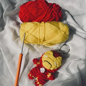 Peut inclure: Un porte-cl&eacute;s personnage crochet&eacute; rouge et jaune avec un masque, un embl&egrave;me de poitrine blanc, des pelotes de laine assorties rouge et jaune, et un crochet en m&eacute;tal avec un manche orange pour les projets manuels.