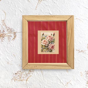 Puede incluir: Un estampado floral enmarcado con una pintura de acuarela de rosas rosas. El estampado está sobre un fondo rojo con un patrón vertical y enmarcado en madera natural. La obra de arte evoca una estética vintage.