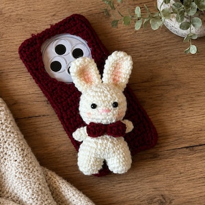 May include: A dark red crocheted phone case featuring a white crocheted rabbit figure attached to the back. The rabbit has black bead eyes, a pink stitched nose, pink-tinted ears, and a dark red crocheted bow tie around its neck.
