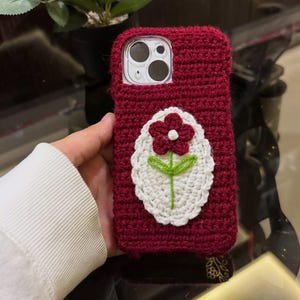 May include: A deep red crochet phone case featuring a central oval patch with a white background. The patch displays a dark red flower with a white center and green stem. The case has a cutout for the camera lens and is made of yarn.