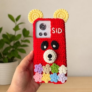 Op de afbeelding: Een rood gehaakt telefoonhoesje met een berengezicht met zwarte ogen, een witte snuit, gele oren en een rij kleine kleurrijke gehaakte bloemen aan de onderkant. De naam SID is in witte draad geborduurd bij de uitsparing voor de camera.