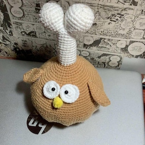 May include: A handmade crochet owl toy in shades of tan, white, and yellow. The owl has large white eyes with black pupils, a yellow beak, and small tan wings. It features a white, cylindrical head with two white spherical shapes on top.