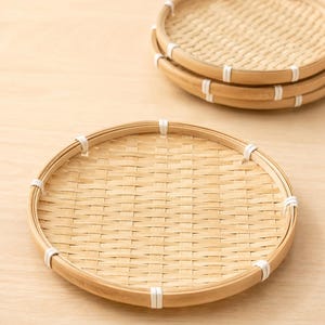 May include: A set of round, woven bamboo trays featuring a light tan natural finish. Each tray has a flat, circular base with a raised rim secured by white cord bindings at regular intervals around the perimeter, creating a structured design.
